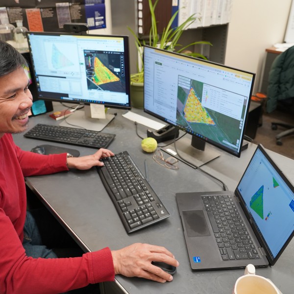 A man works on three computers. He is wearing a red shirt, sitting at a desk and smiling.