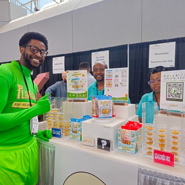 Three people, one of whom is wearing a green body suit, standing at a trade show booth. 
