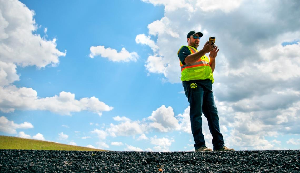 A man standing in the middle of a road talking and looking at a small device in his hand with blue skies behind him 