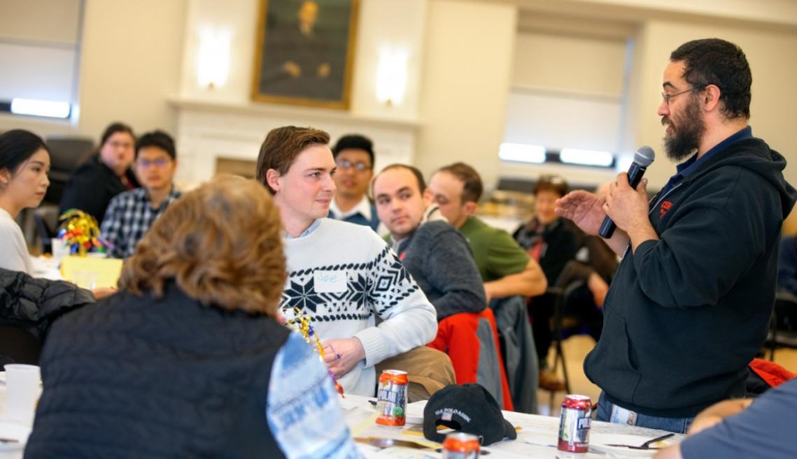 A man standing and talking into a microphone in front of a room full of people sitting at tables listening to him