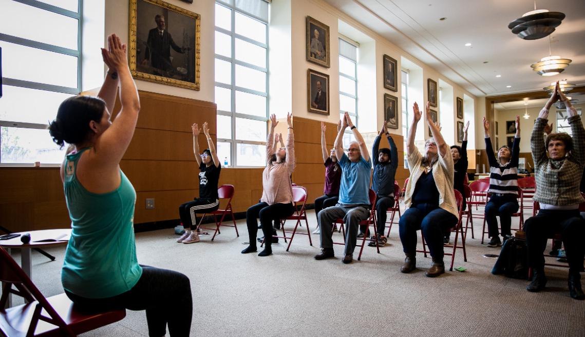 A group of males and females sitting in chairs inside following a chair yoga instructor 