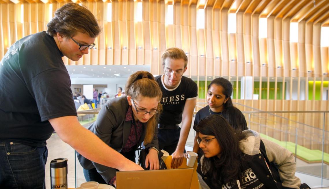 Two men and three females standing and working around a computer