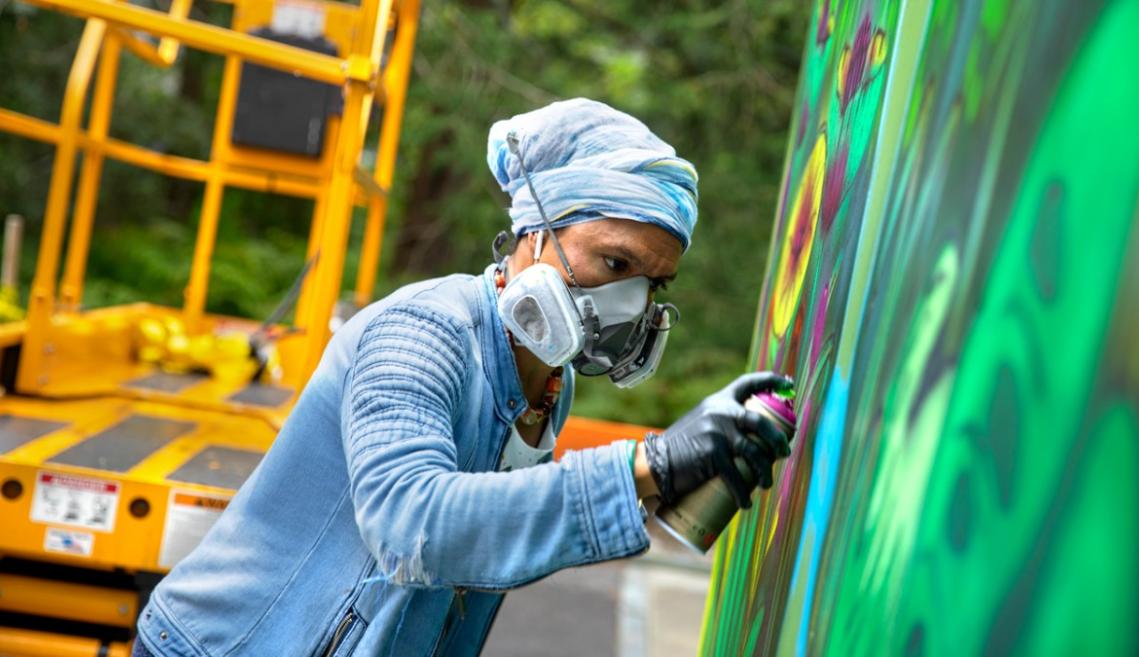 A masked man spraypaints a wall, creating a mural