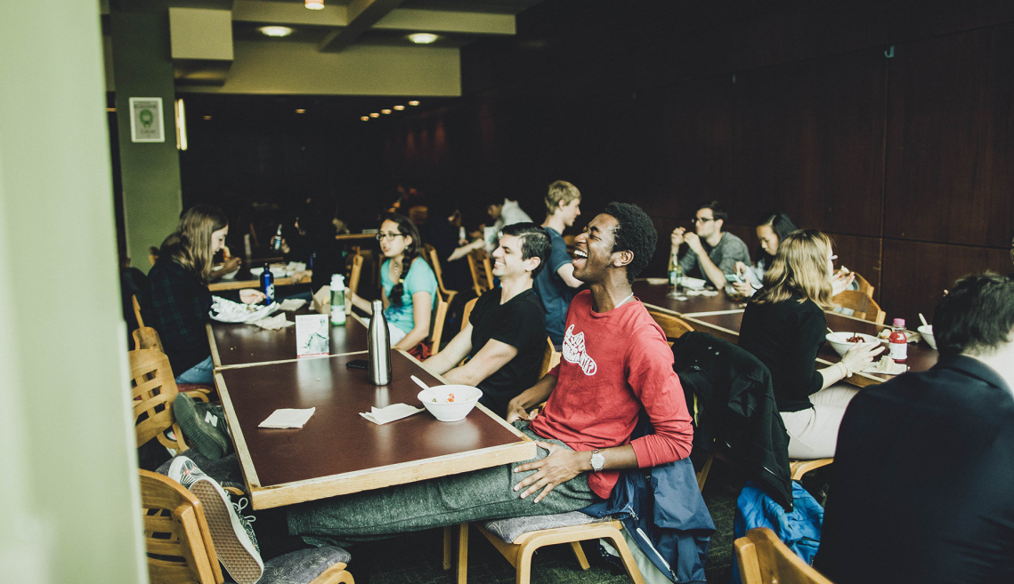 students sit at tables in a dining hall and some of them are laughing