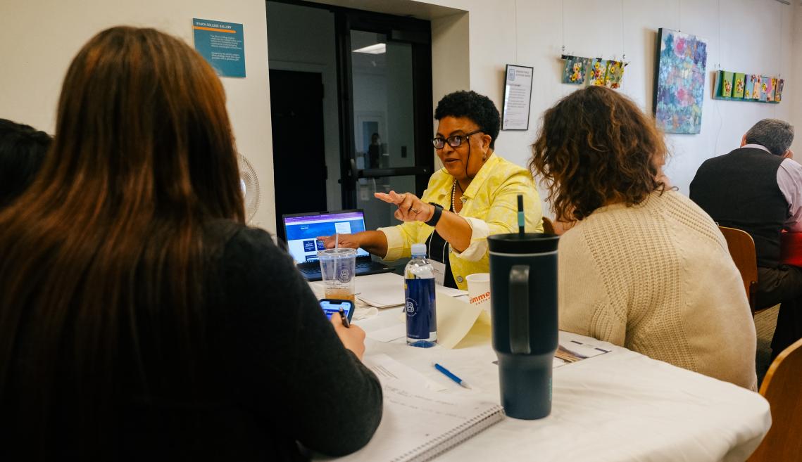 Photograph of a few people sitting looking at a presentation on a laptop screen while one woman in a yellow shirt is talking toward them.