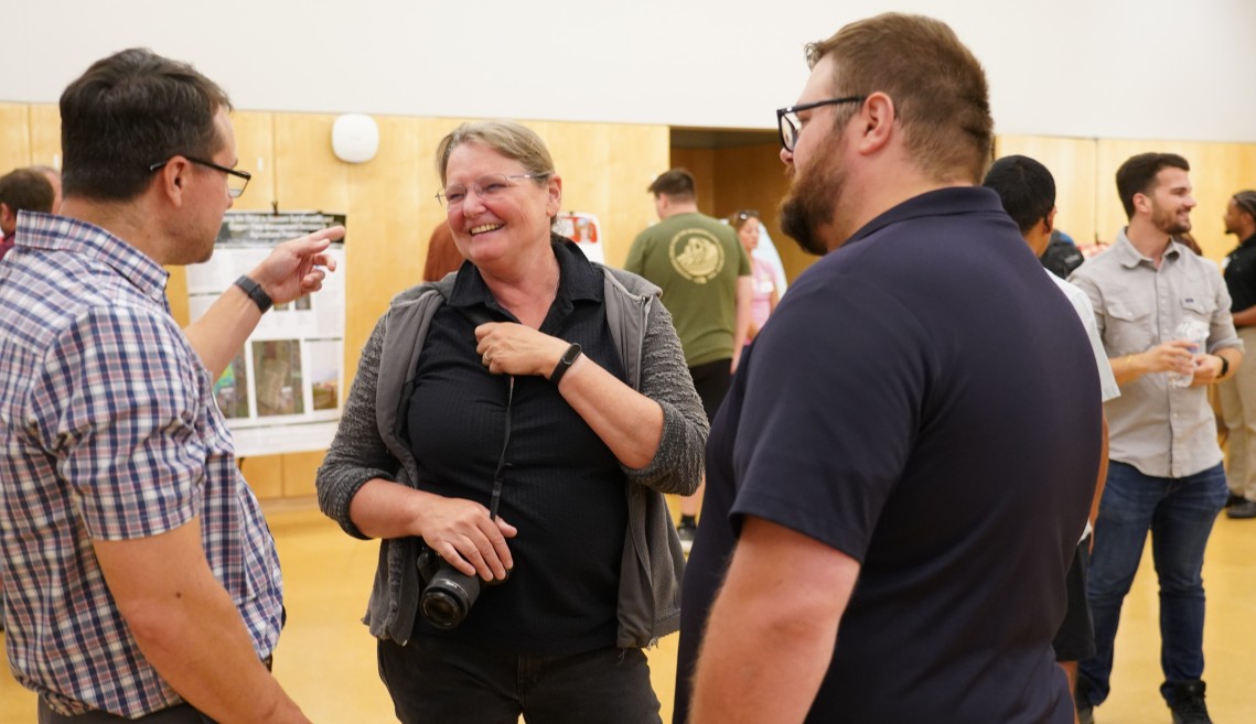 A woman facing toward the camera smiles while two men talk to her.
