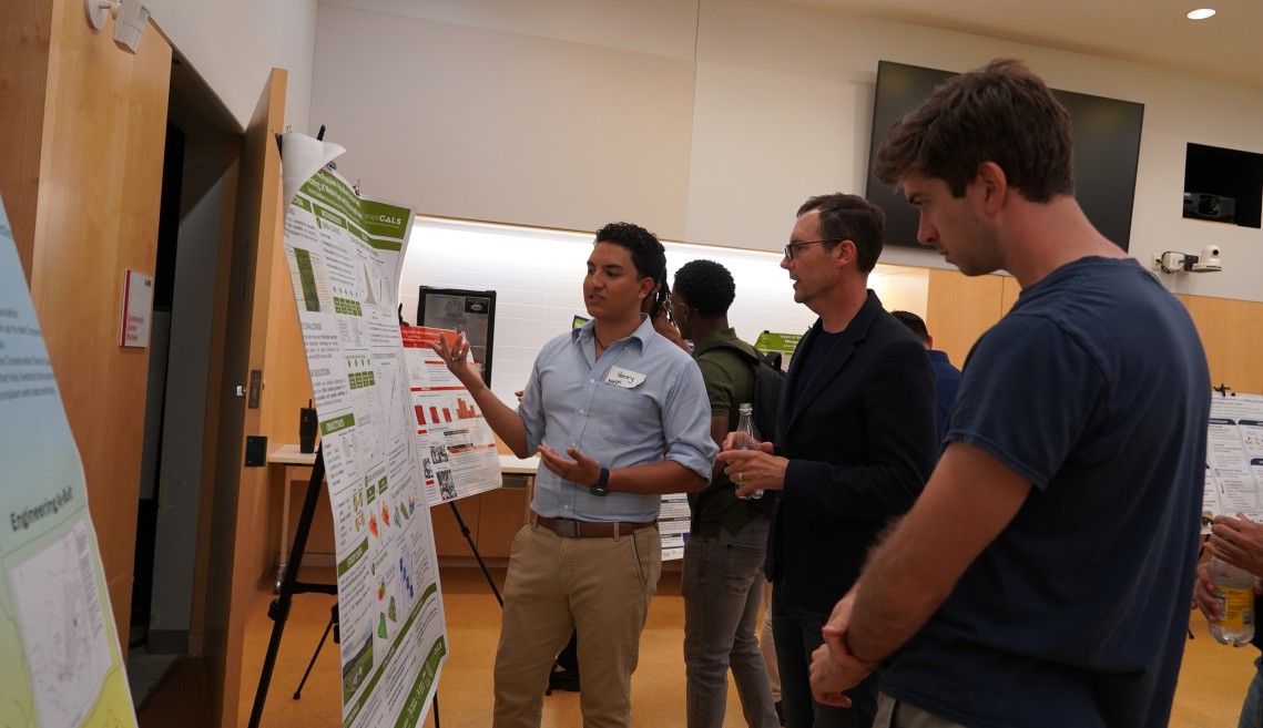 A man presents his research to two other man, gesturing toward his research poster on his left.