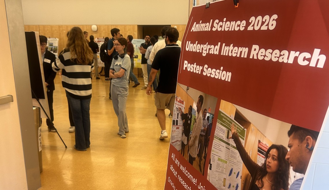 An introductory poster sits on an easel outside of the conference room where the poster session was held. Multiple people stand in that room talking to one another.