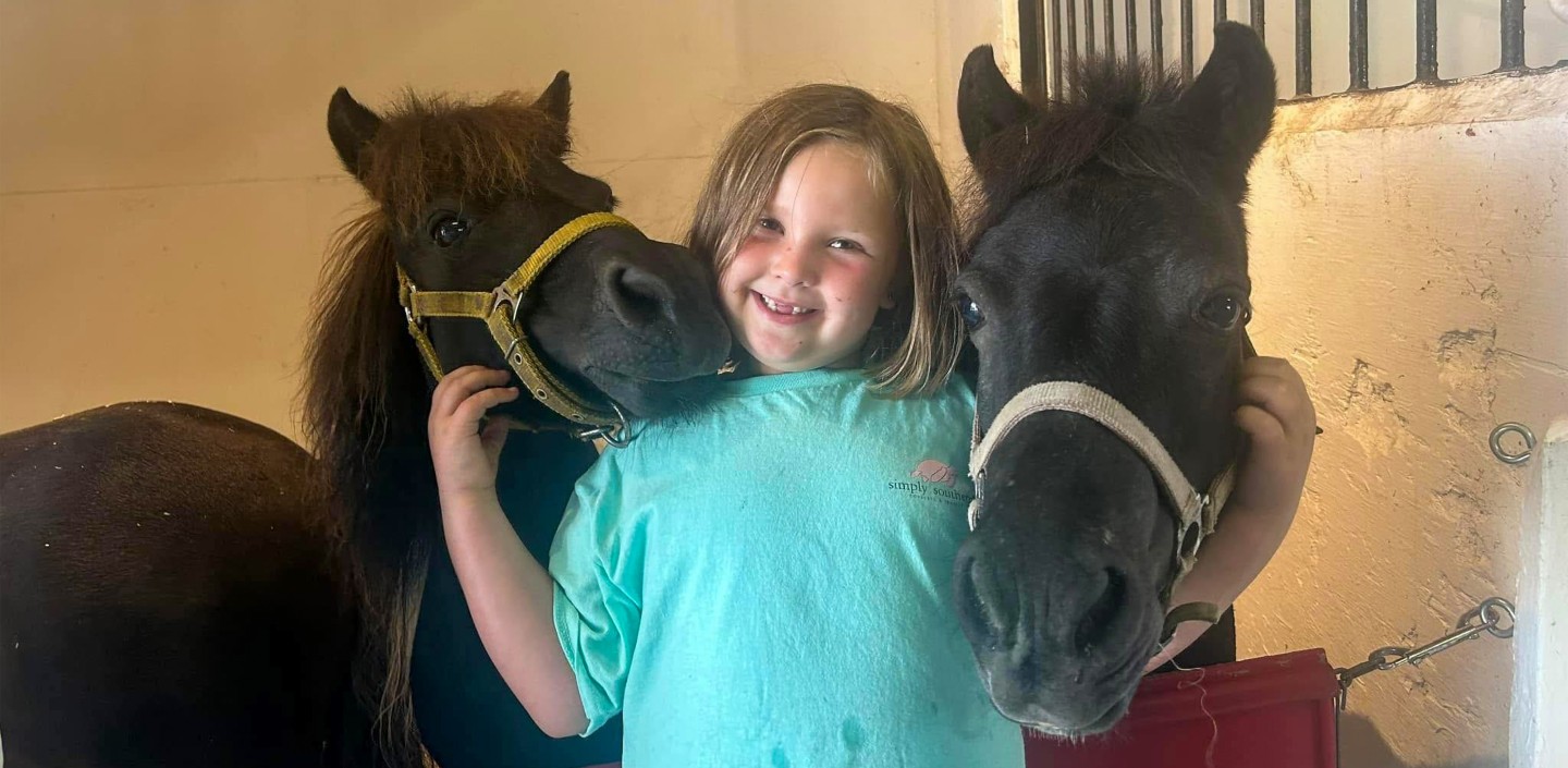 a young girl stands between two ponies with her arms cradling their heads