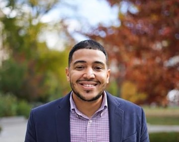 portrait of a man in a jacket and pink shirt in front of trees on the quad