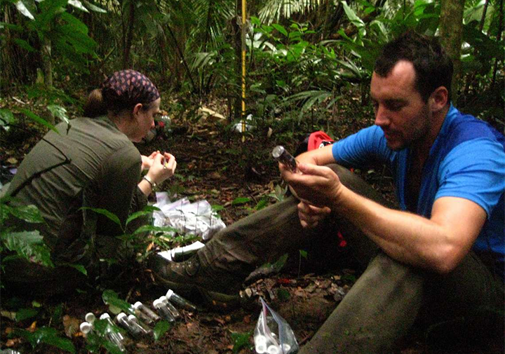a man and a woman sit on the rainforest floor holding vials