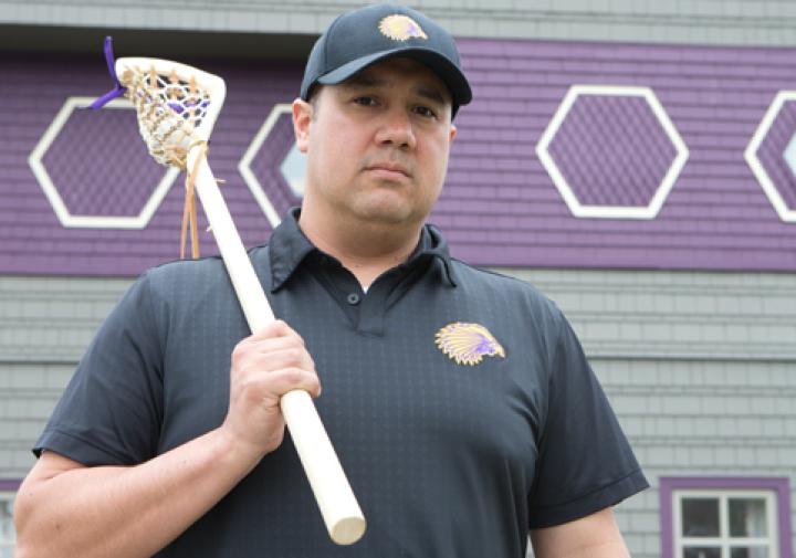 Picture of man holding a lacrosse stick in front of the side of a grey and purple building.