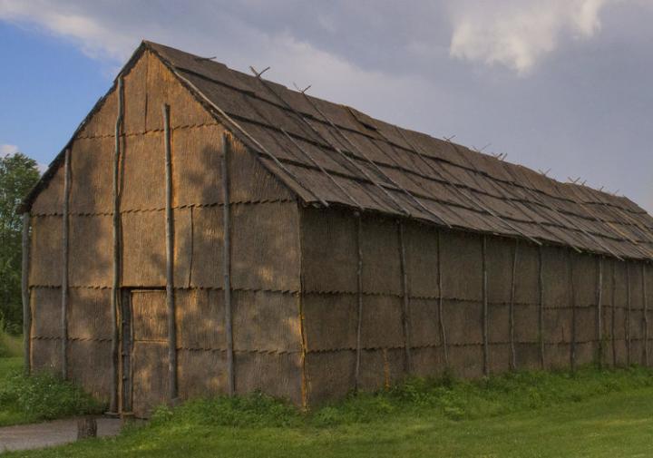 Picture of a traditionally built longhouse in a field with a rainbow behind it in the sky.
