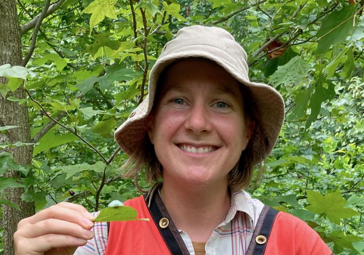 person with a hat and vest in the woods smiling and holding up a leaf with something on it