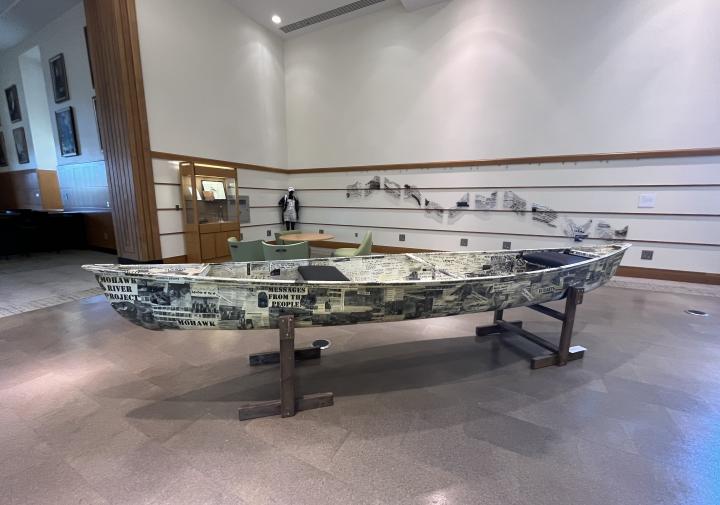 Photograph of a canoe in an exhibit covered with black and white photos and hand written messages.