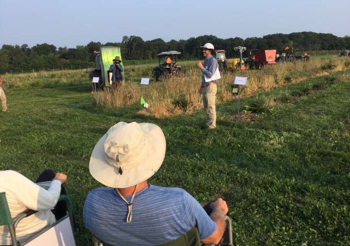Several people are sitting in the foreground listening to a speaker present research in a Christmas tree field with farm equipment in the background.