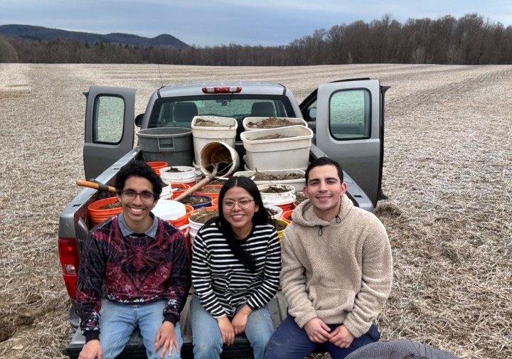 Three people sit next to each other in a truck bed. One man is at the front taking a selfie of everyone.