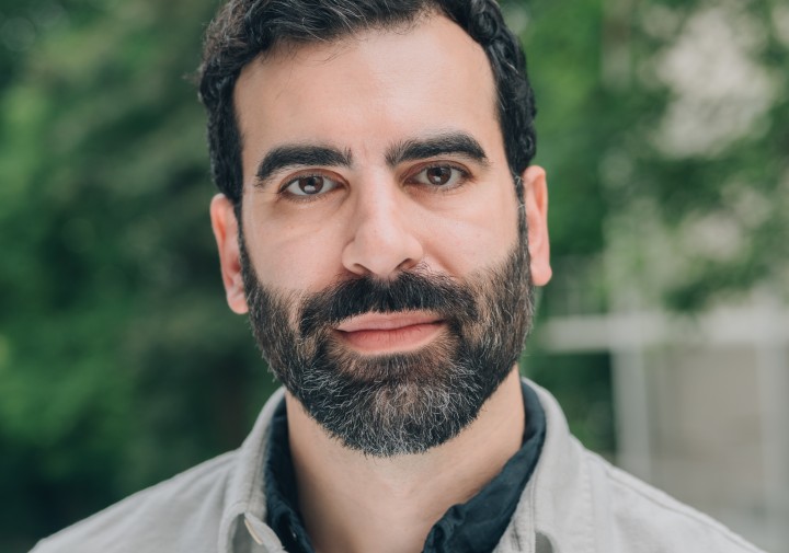 Bearded man wearing a light button-down shirt, standing outdoors with trees softly blurred in the background.