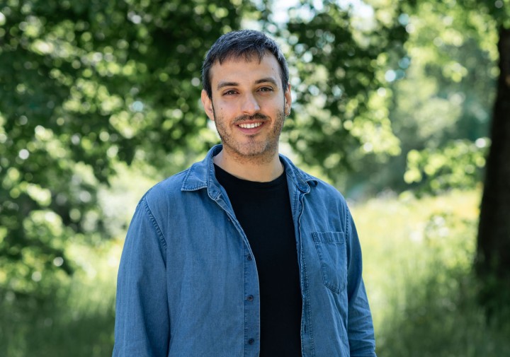A headshot image of Joan Casanelles Abella - Joan is wearing a black shirt under an unbuttoned blue button-down shirt while standing in a shady clearing smiling at the camera.