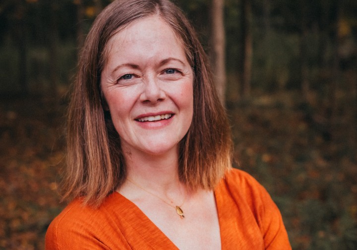 Professional portrait of person standing with crossed arms and long brown hair wearing an orange shirt.