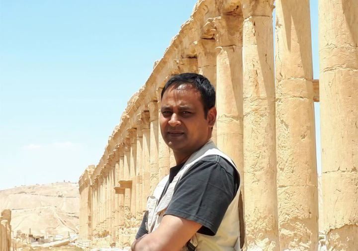 Man standing with arms crossed beside a row of ancient stone columns in a desert setting.