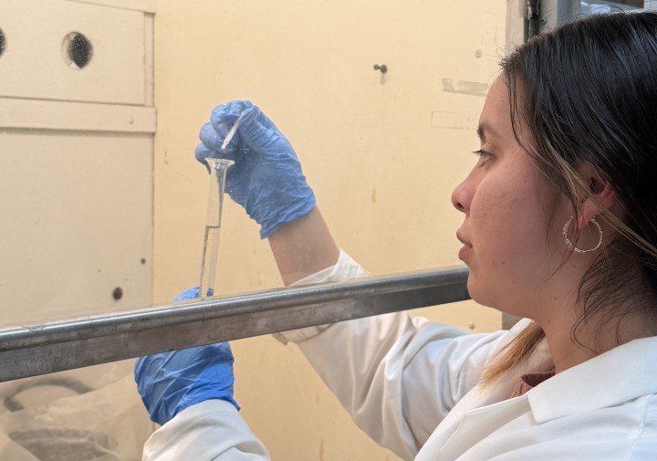 A woman deposits a substance into a test tube under a fume hood.