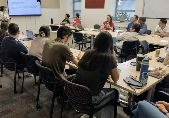 A bunch of people sitting at tables. The tables are roughly in a circle. The people watch a screen and presentation at the front of the room. 
