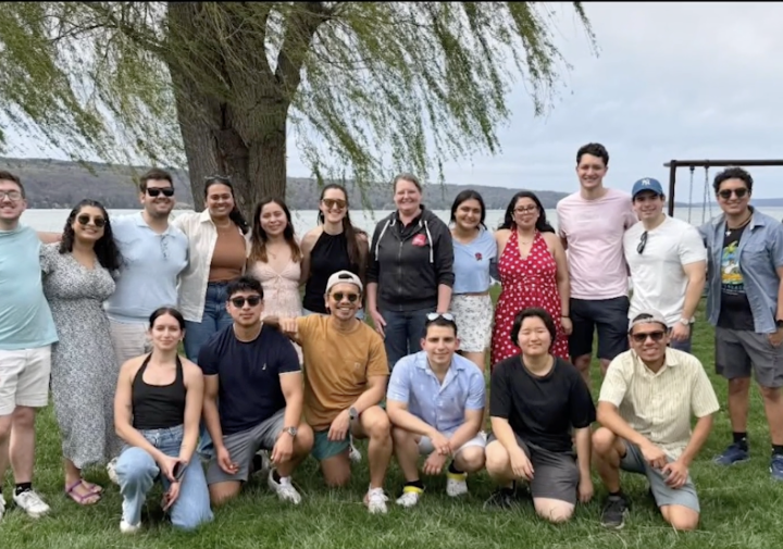A group smiles with their arms around each other, with a lake and a tree in the background.