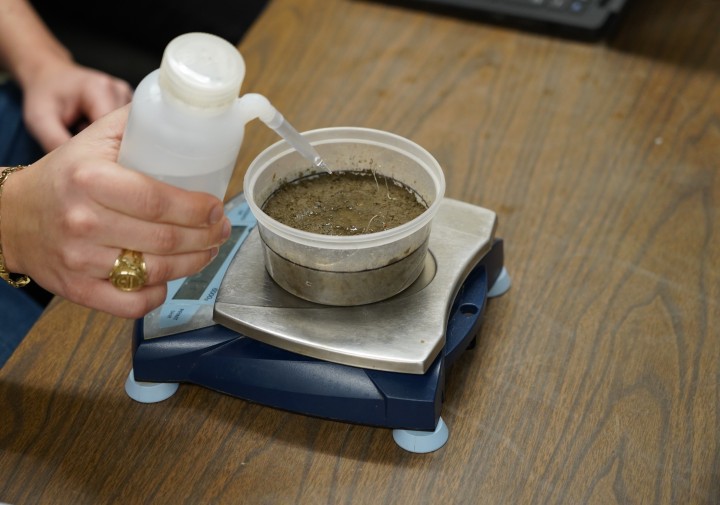 A soil sample sits on a scale. A hand is squeezing water out of a bottle onto the sample.