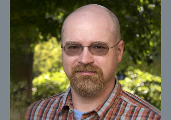 A headshot of a person with glasses and a light-colored goatee standing in front of a sun-dappled grove