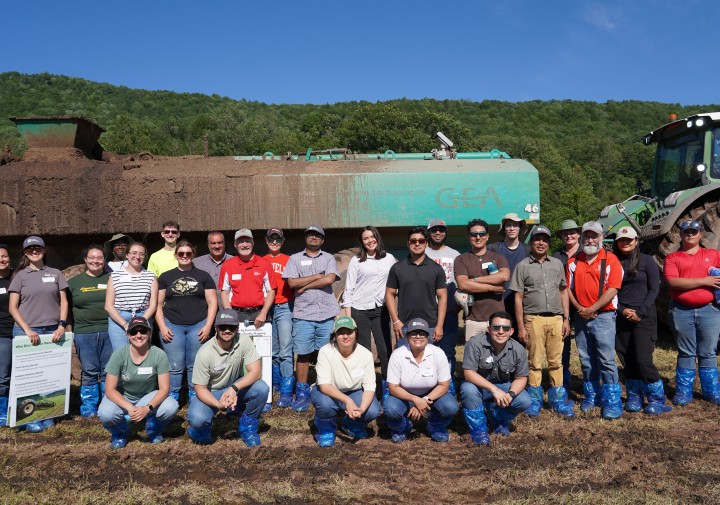 A group of people stands in front of a manure injector in a field, smiling.