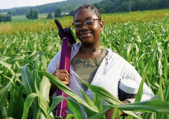 A woman stands in a corn field holding a piece of equipment.