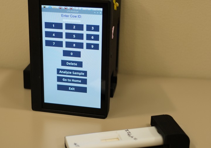 a small square piece of tech with a touch screen sits on a table with a plastic test strip in front of it