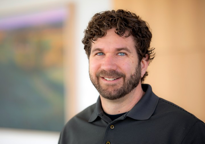 A head-and-shoulders portrait of a smiling man looking directly at the camera. He has curly, dark brown hair, a full beard and mustache, and bright blue eyes. He is wearing a dark gray, short-sleeved polo shirt with a fine, horizontally ribbed texture. The background is blurred, showing a framed artwork with soft, abstract colors on the left and a plain, light wood-toned surface on the right.
