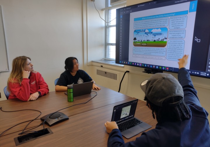Two women sit at a table on the left, one man sits on the other side of the same table on the right. They are all looking towards a screen in the center.