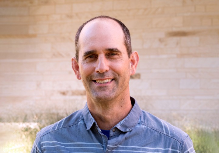 A portrait of a middle-aged man smiling directly at the camera. He has short dark hair on the sides of his head, and light stubble. He is wearing a blue button-down shirt with thin horizontal white stripes. The background is an out-of-focus light tan stone wall with some blurred green vegetation at the bottom.