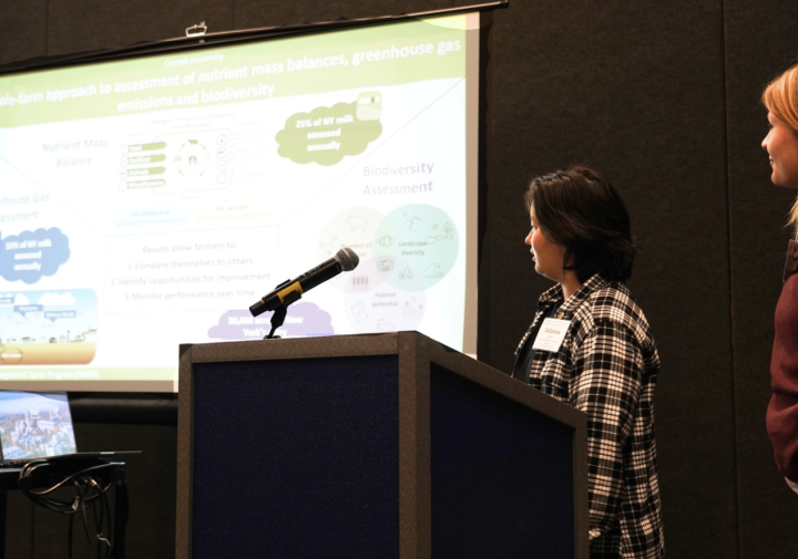 Two women stand to the left of a slideshow presentation in front of a dark background.