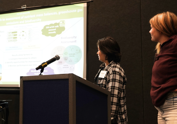 Two women stand to the left of a slideshow presentation in front of a dark background.