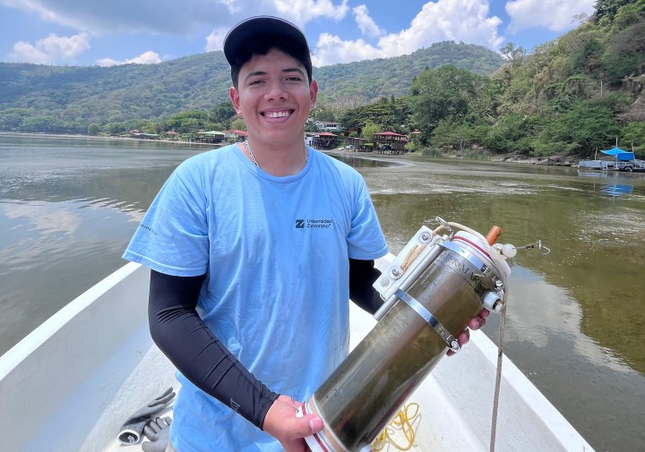 A man stands in a floating boat and holds a jar of dirty water.