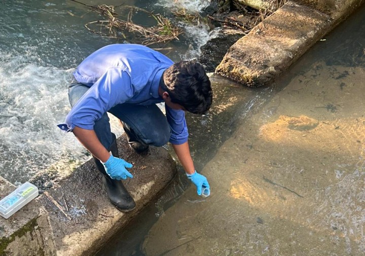 A man wearing gloves kneels next to a water body and dips a small container into it.