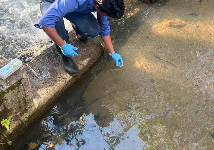 A man wearing gloves kneels next to a water body and dips a small container into it.
