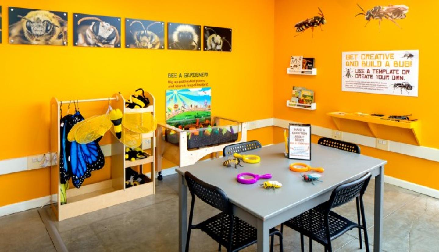 A yellow room is filled with images of bees and a table with magnifying glasses and plush bugs