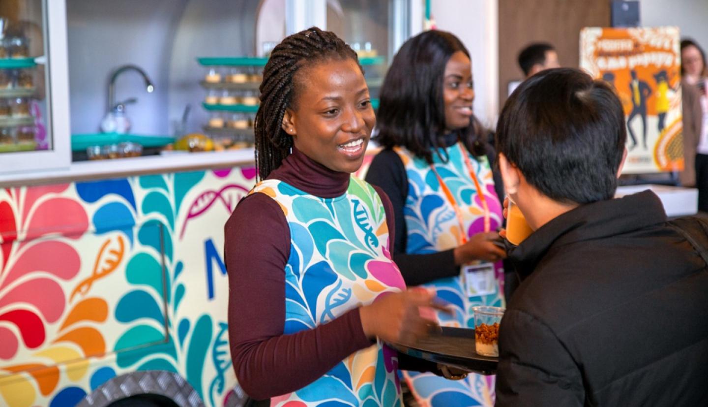 Two women standing in front of a colorful food truck wearing matching colorful clothing serving drinks on trays