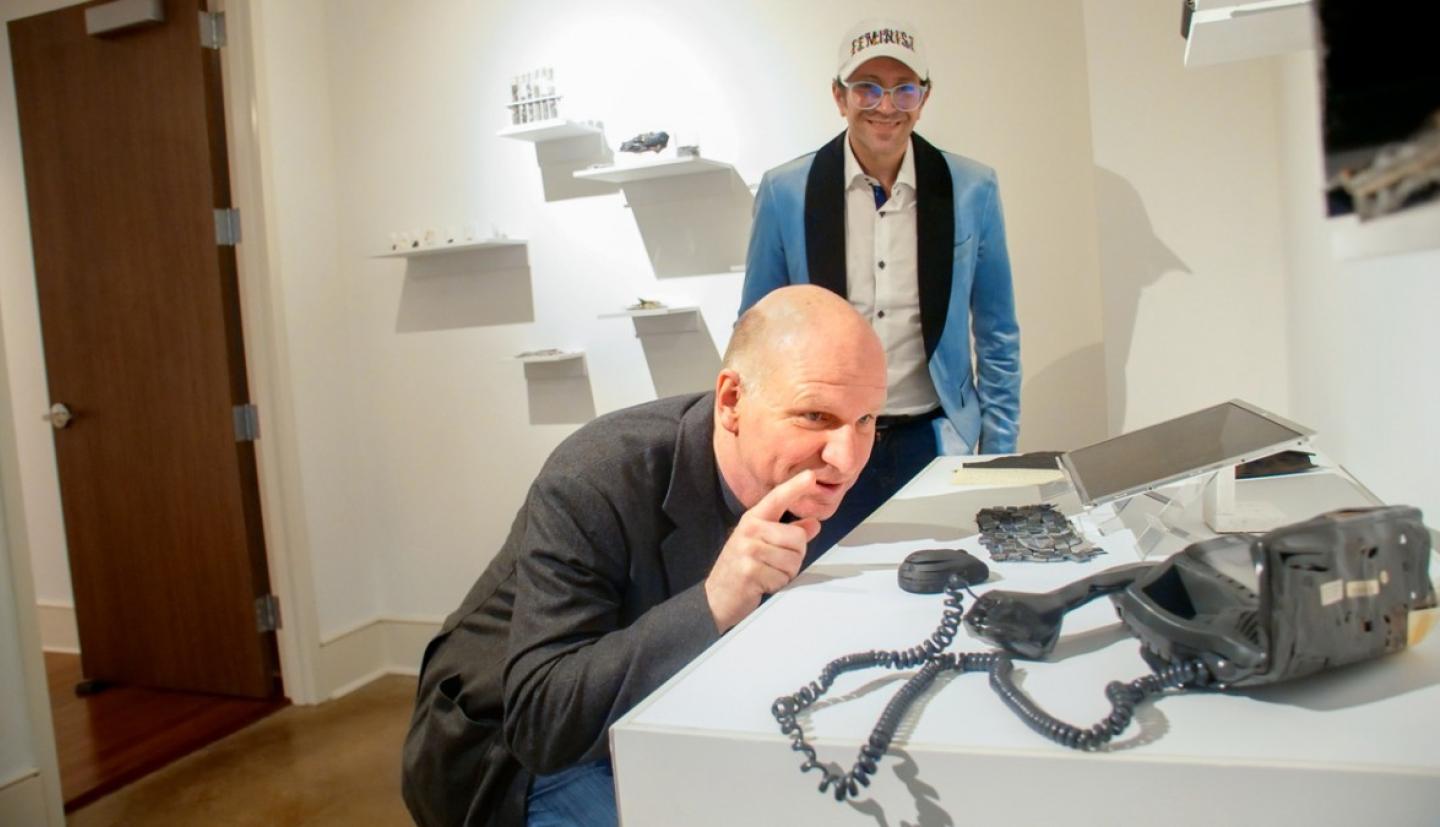 A man crouching at a table looking at art while another man looks on