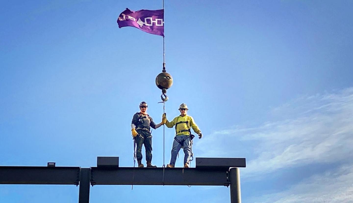 Two men standing on a building structure holding a cloth image used to represent the  the sovereignty of the Six Nations of the Haudenosaunee Confederacy