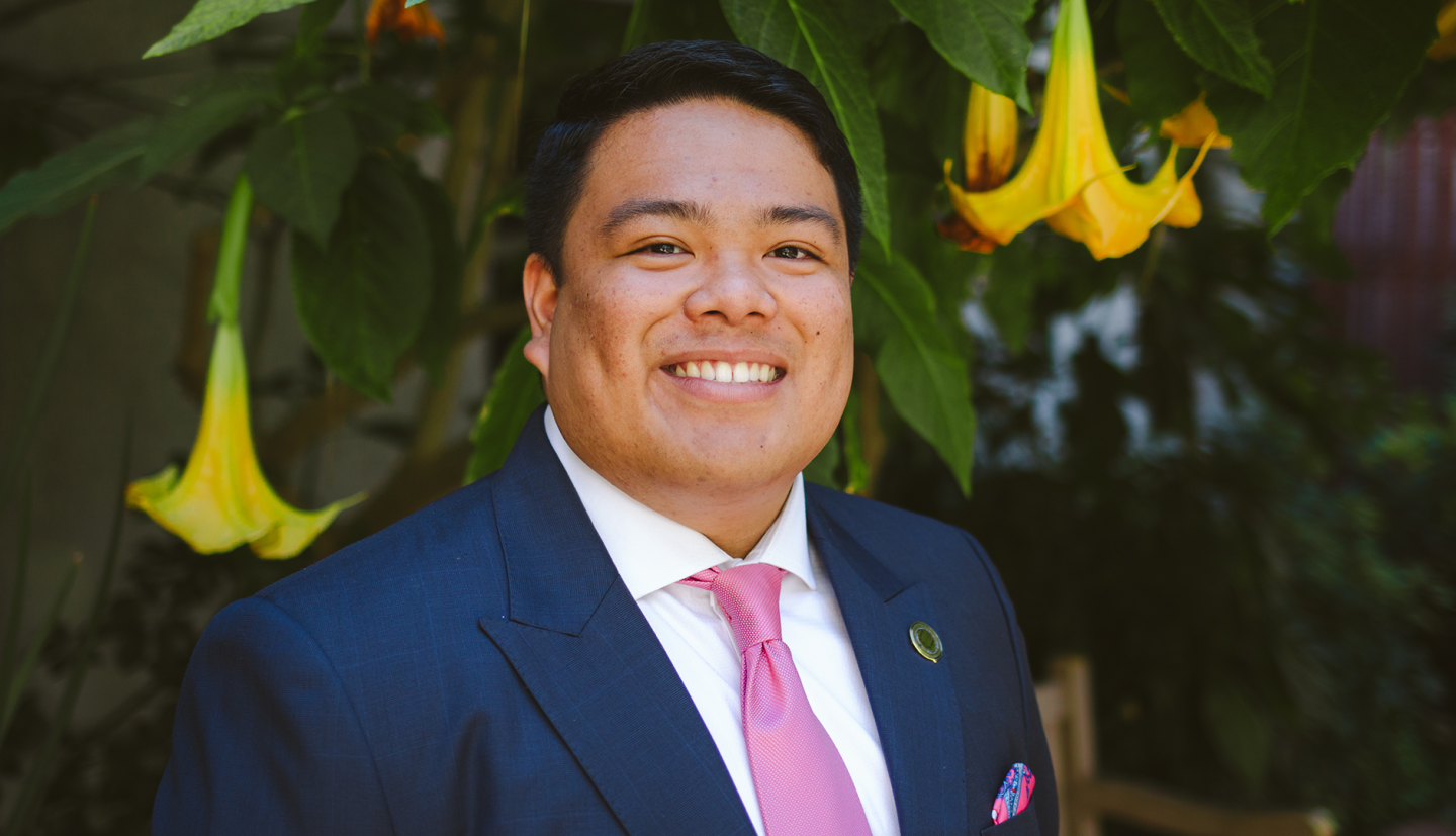 a student in a blue suit and pink tie stands in front of flowers