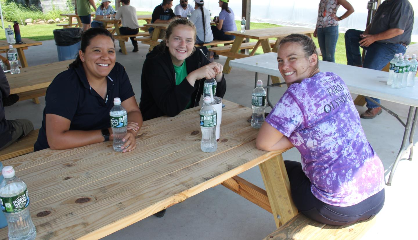 Carly sitting at a picnic table, turned to face the camera smiling. Two other people are on the other side of the table, also smiling.
