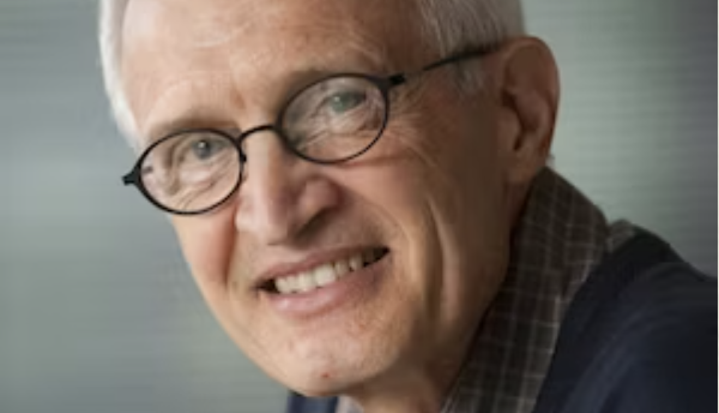 A man with white hair and oval glasses smiles at the camera with his head slightly turned. He is wearing a checkered shirt. 