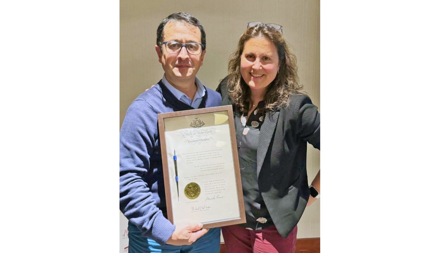 Alejandro and Julie standing next to each other holding a framed document with a pen attached to it between them.