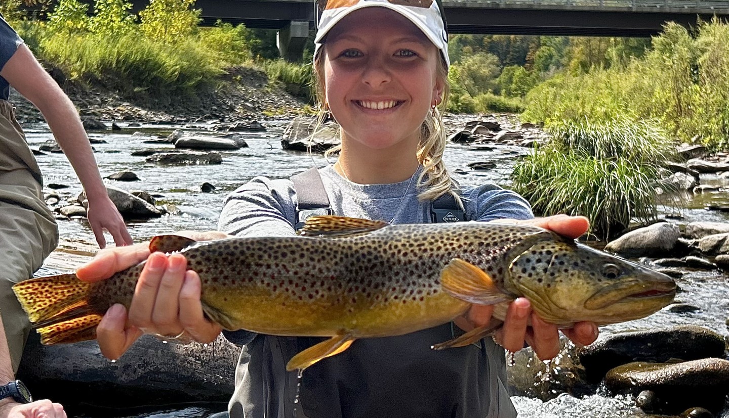 Photograph of a woman wearing a hat holding a trout in a stream with green tree covered mountains behind.